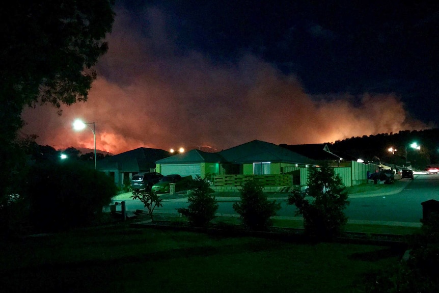 Thick smoke and an orange glow from a bushfire looms over the horizon and rooftops of houses at night.