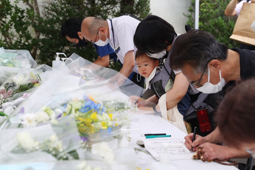 Asian people wearing masks stand side-by-side as they write notes on tables covered with flowers 