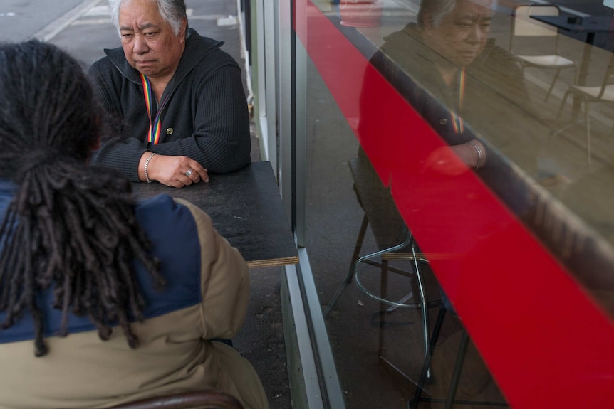 Maggie sits at sidwalk table with Scott, her image reflected in the cafe window.