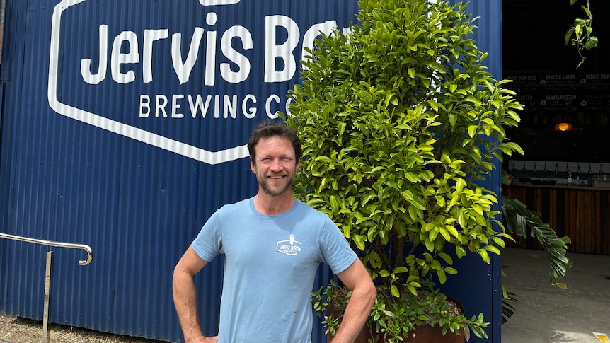 A smiling, dark-haired man with a neat beard stands with his hands on his hips outside a warehouse.