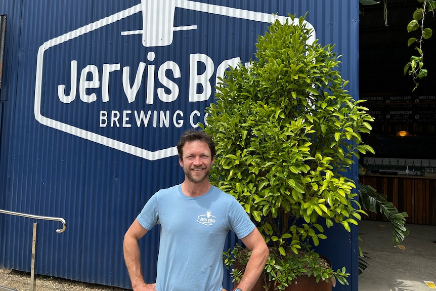 A smiling, dark-haired man with a neat beard stands with his hands on his hips outside a warehouse.