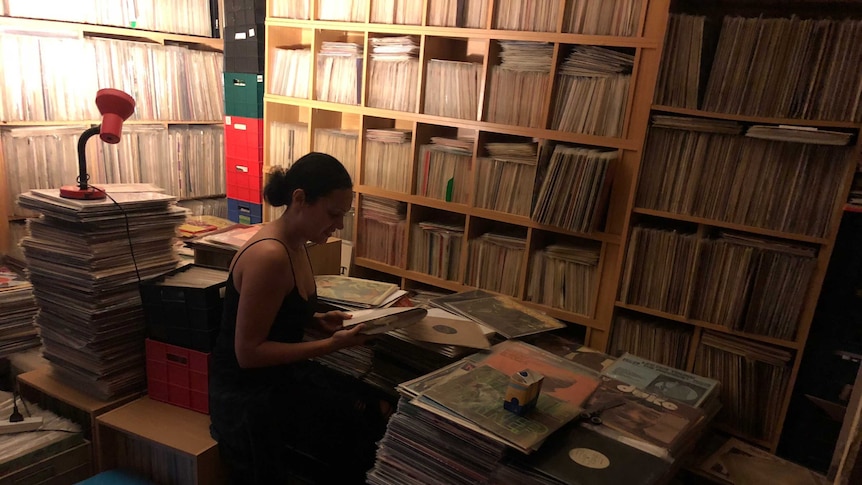 Records are stacked in shelves from floor to ceiling, the collection is also in boxes on the floor. Natalie sits between them.
