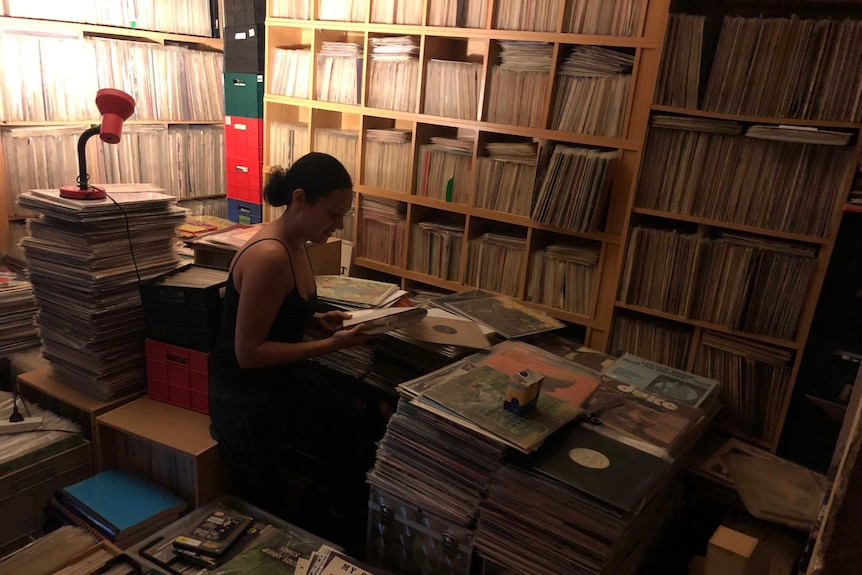 Records are stacked in shelves from floor to ceiling, the collection is also in boxes on the floor. Natalie sits between them.