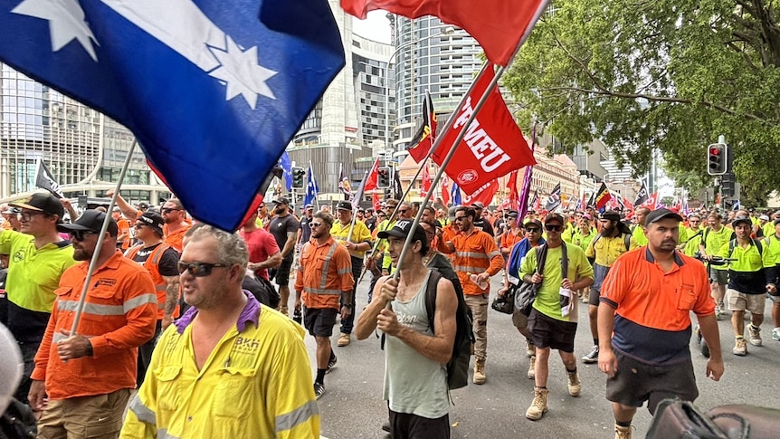 An image of construction workers in high vis workwear marching with flags in Brisbane outside parliament