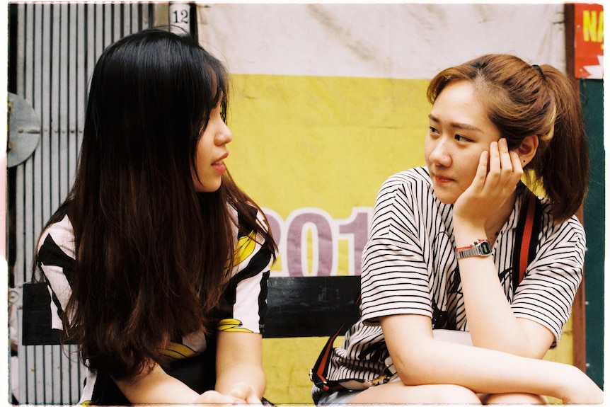 Two Asian women sitting and looking at each other. One is resting her face on her hand