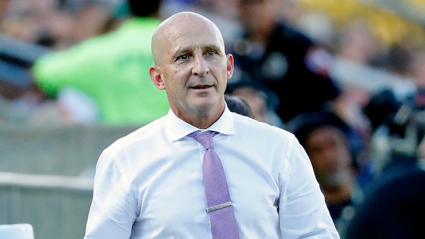 A man with bald head wearing white business shirt on a playing field on a sunny day