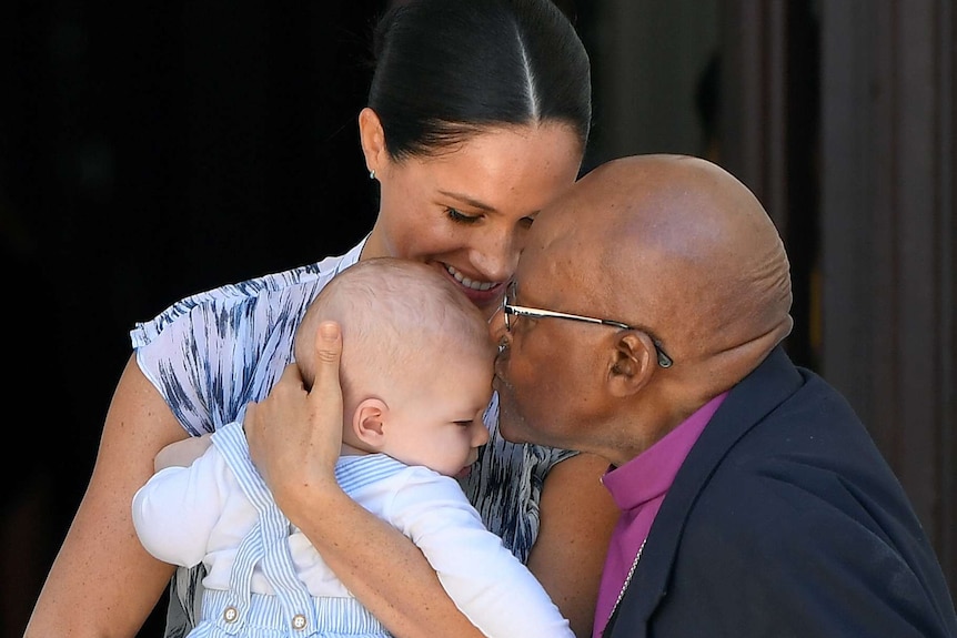 Meghan smiles while holding Archie. Archbishop Tutu leans in to kiss Archie on the forehead.