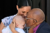 Meghan smiles while holding Archie. Archbishop Tutu leans in to kiss Archie on the forehead.