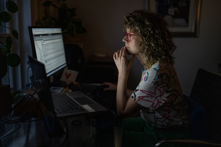 A woman wearing glasses sits looking at a computer in a darkened room. Her hand is resting on her chin.