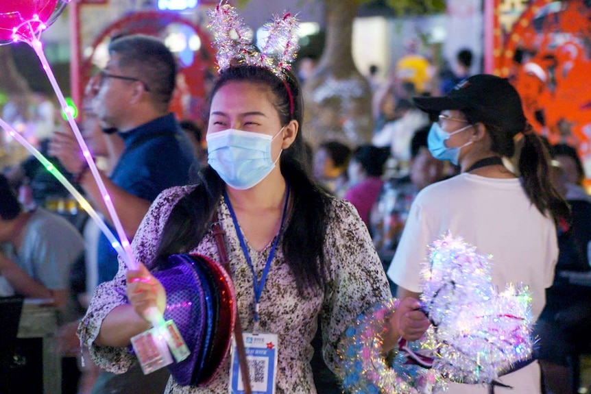 A woman in a face mask holds bunny ears and light-up balloons for sale