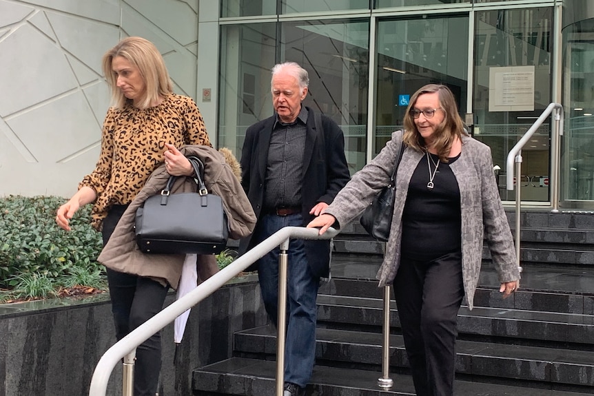 Two middle-aged women walk down stairs outside the Perth District Court building with an older man between them.