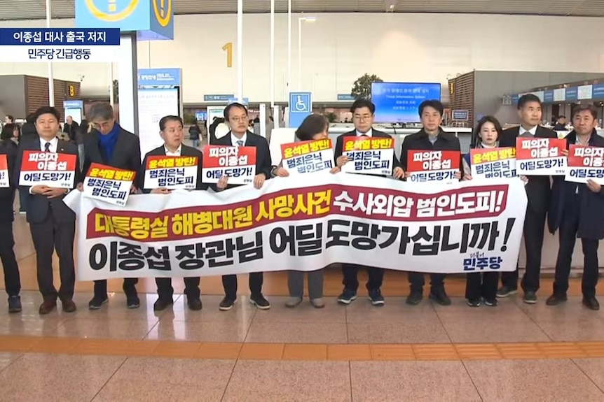 A row of people holding signs and a banner stand in an airport terminal.