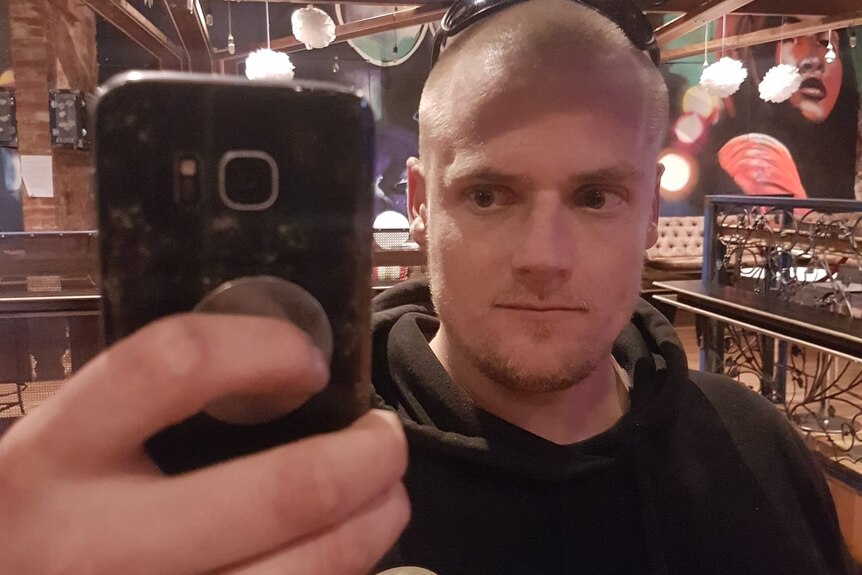 A man with a shaved head takes a photo of himself on his phone through a mirror