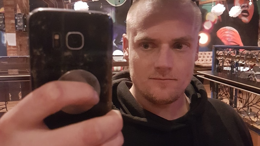 A man with a shaved head takes a photo of himself on his phone through a mirror