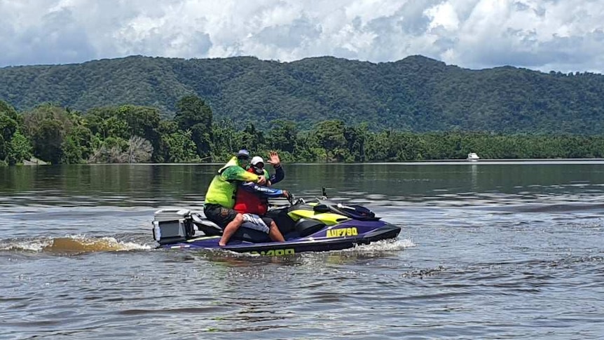 Two people sit on a jet ski in a river waving at the camera