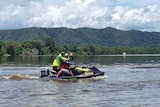 Two people sit on a jet ski in a river waving at the camera