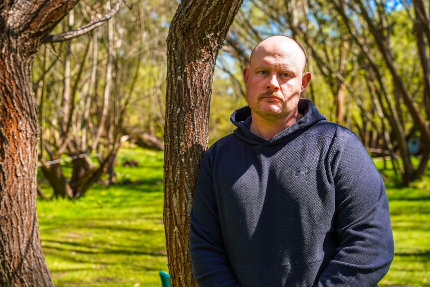 A man wearing a black hoodie and jeans leans against a tree.