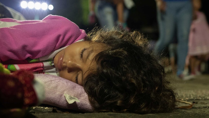A young girl with wavy brown hair and wearing a pink top sleeps on the footpath with people walking past in the background