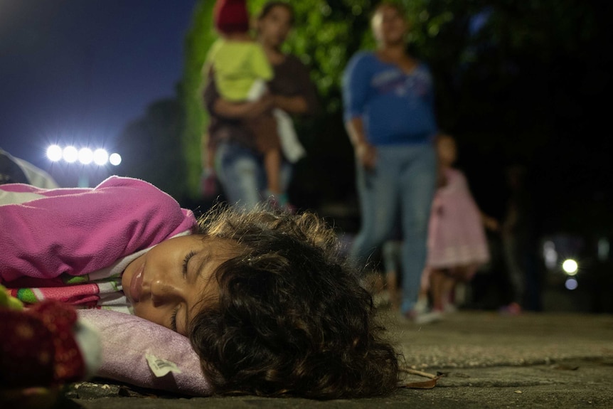 A young girl with wavy brown hair and wearing a pink top sleeps on the footpath with people walking past in the background