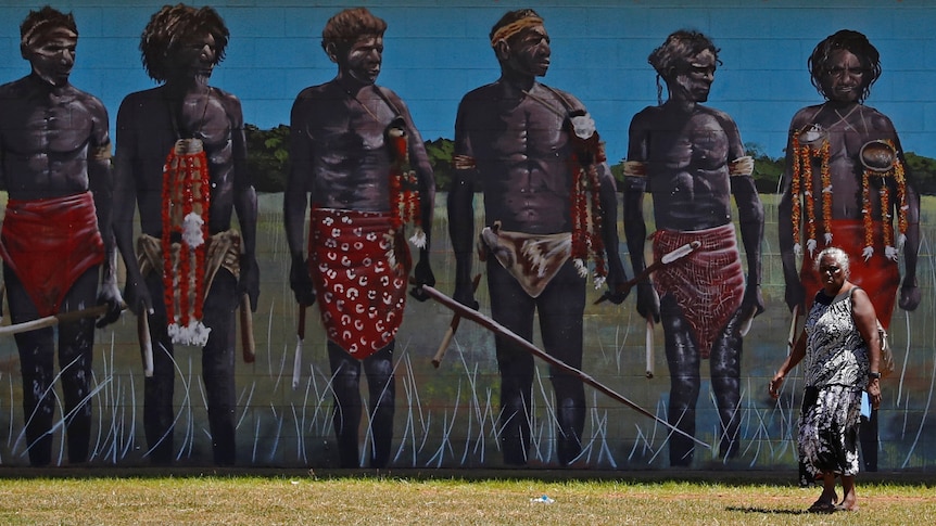 An Indigenous woman walks past a mural of Indigenous people wearing traditional dress.