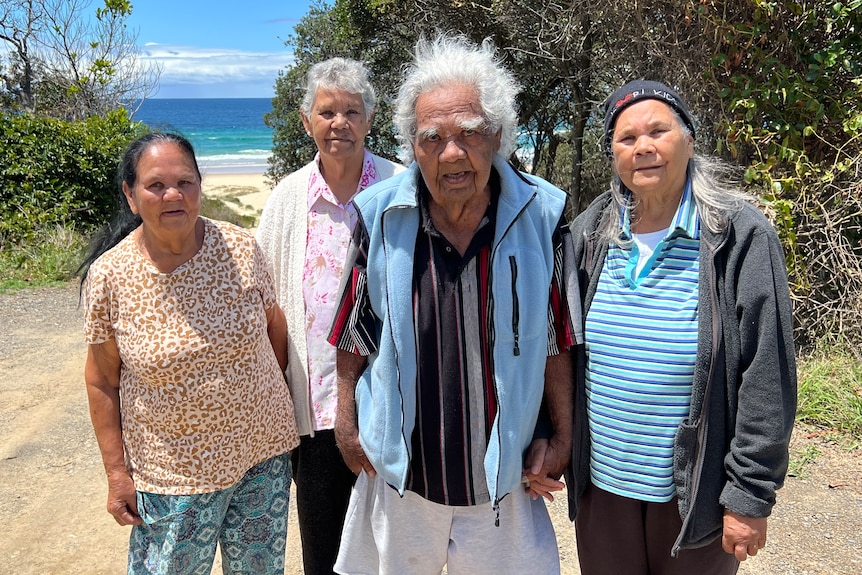 Elderly Aboriginal man and three elderly Aboriginal women stand slightly behind him at a bush and beach location, not smiling. 