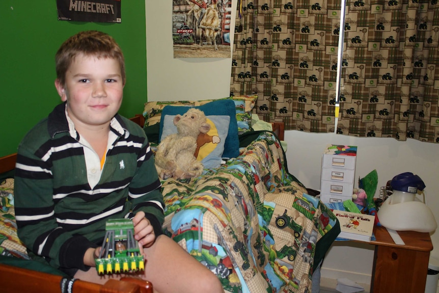 Nine-year-old Clancy Stretton sits on his bed with a tractor-print bedspread, holding a toy cotton harvester toy.