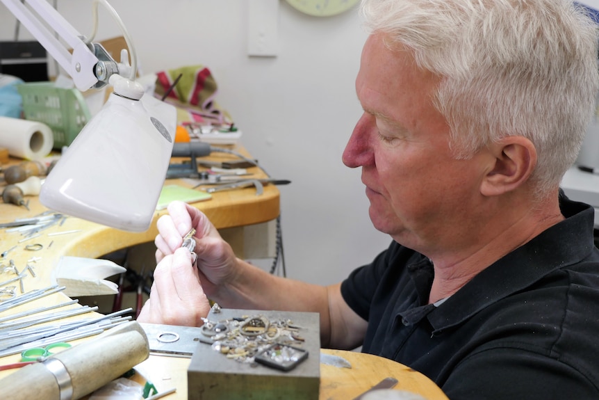 Un hombre con canas sostiene un anillo y lo mira en su taller de joyería.