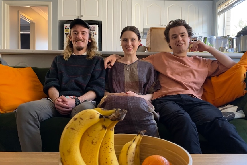 Three people sit on a couch with a bowl of fruit in the foreground