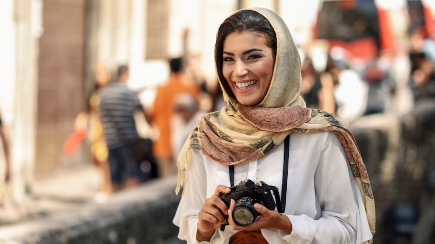 A smiling woman wearing a hijab holds a camera while standing on a busy street