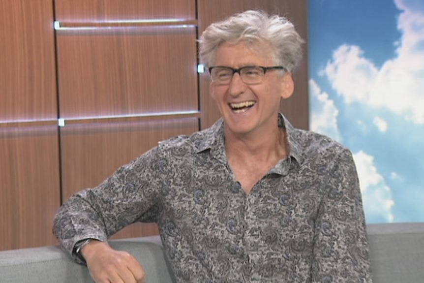 A man laughs while sitting on the set of a television news show.