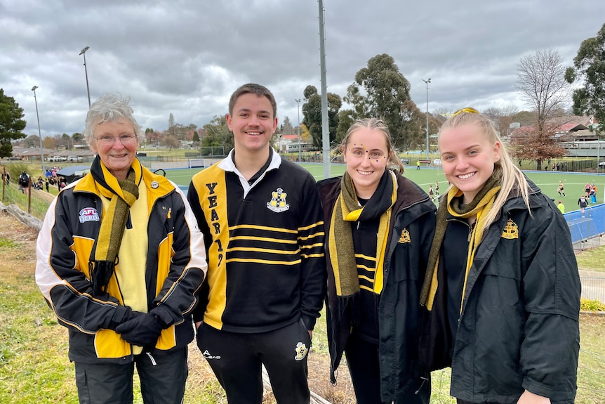 An older woman standing with three high school students, all wear yellow and black team colours.