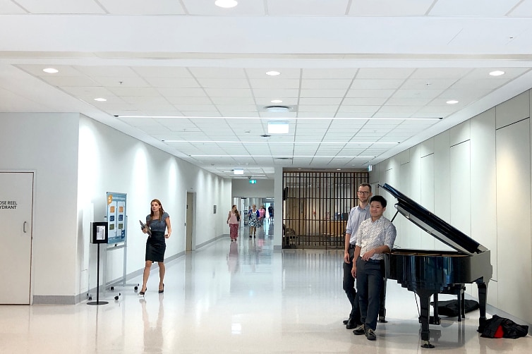 Grand piano in the foyer of a large hospital with two men standing beside it smiling and people walking in the background.