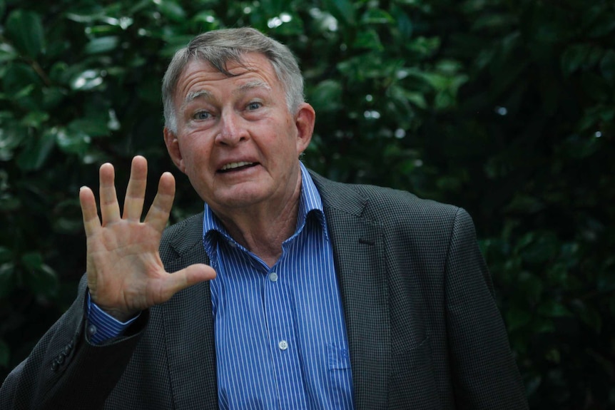 Veteran swimming coach  Laurie Lawrence waving in front of a hedge.