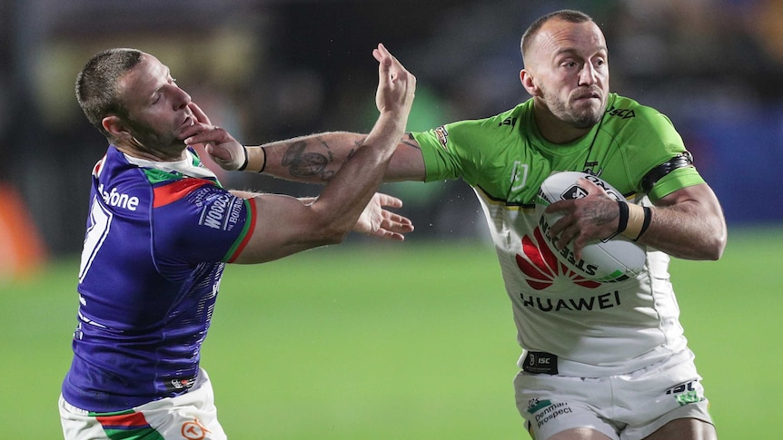 A male NRL player holds the ball with his left hand as he uses his right arm to palm off an opponent.