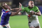 A male NRL player holds the ball with his left hand as he uses his right arm to palm off an opponent.