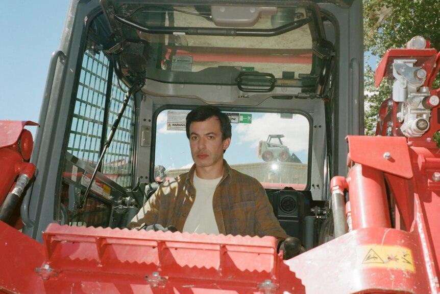 Nathan sits in a big red tractor with a bewildered expression, wearing a shirt over a white tee.