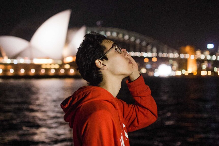 A young man wearing a red jumper is looking up and touching his glasses. It is night and the Sydney Opera House is behind him.