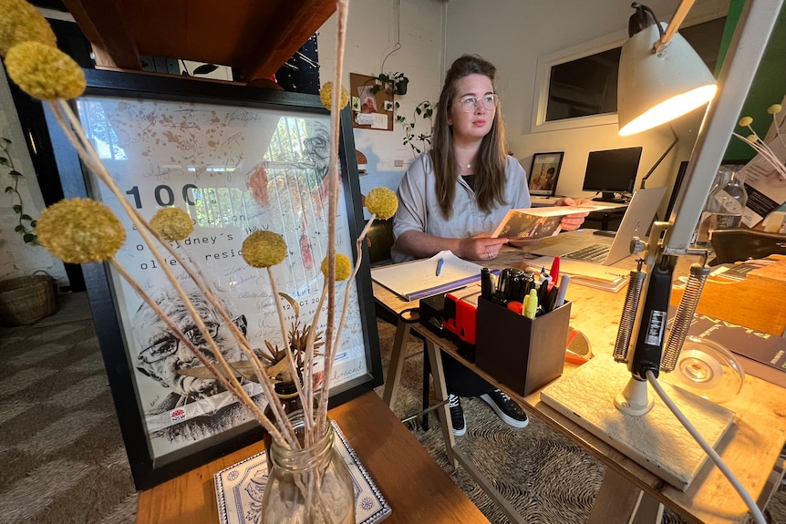 A woman wearing glasses sits in an art studio. 