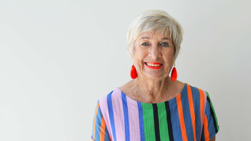 An elderly lady standing in front of a white wall dressed in a multi-coloured dress.