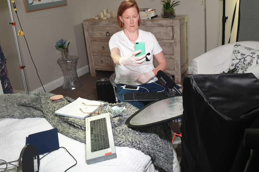 Sales holding mobile phone in front of face surrounded by camera and lights in home studio in her bedroom.