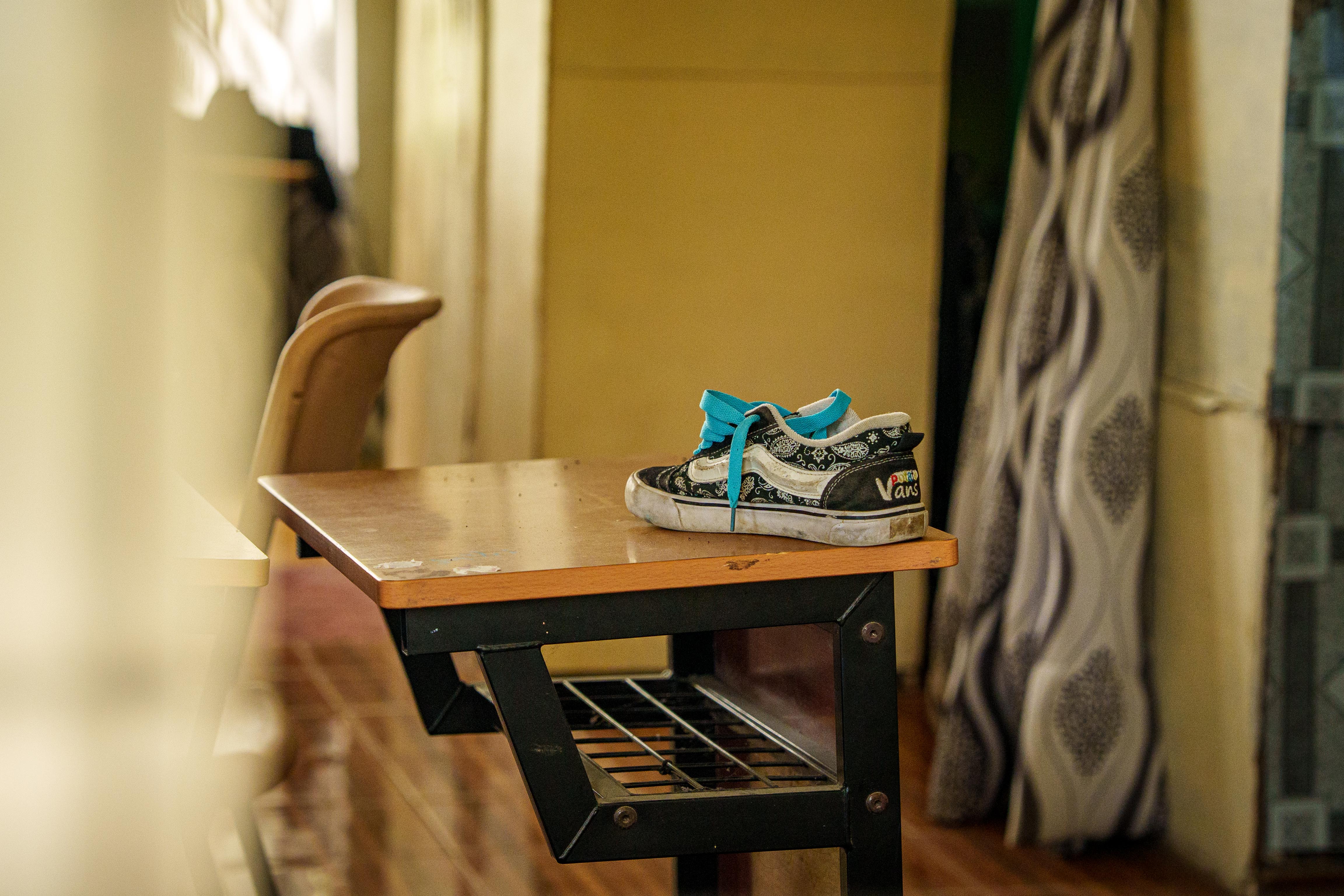 A single black and white Vans sneaker with blue laces sits on top of a wooden desk inside a classroom.