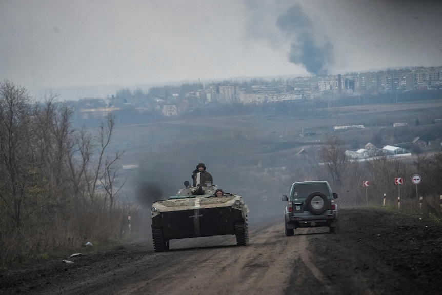 Soldiers ride on a armoured vehicle down a dirt road away from a settlment where smoke is rising into a grey sky.