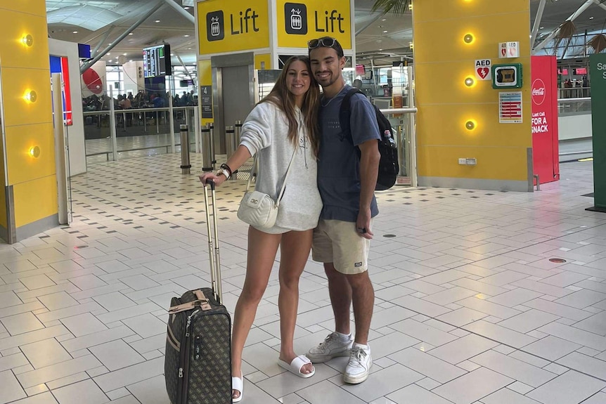 Woman in white hoodie and shorts with small suitcase, with man in t-shirt and shorts at airport terminal, both smiling