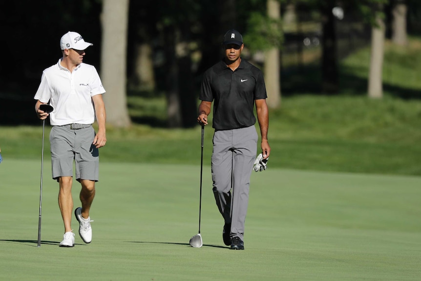 Tiger Woods, in a black shirt grey trousers, walks next to Justin Thomas, in a white shirt and grey shorts on a golf course