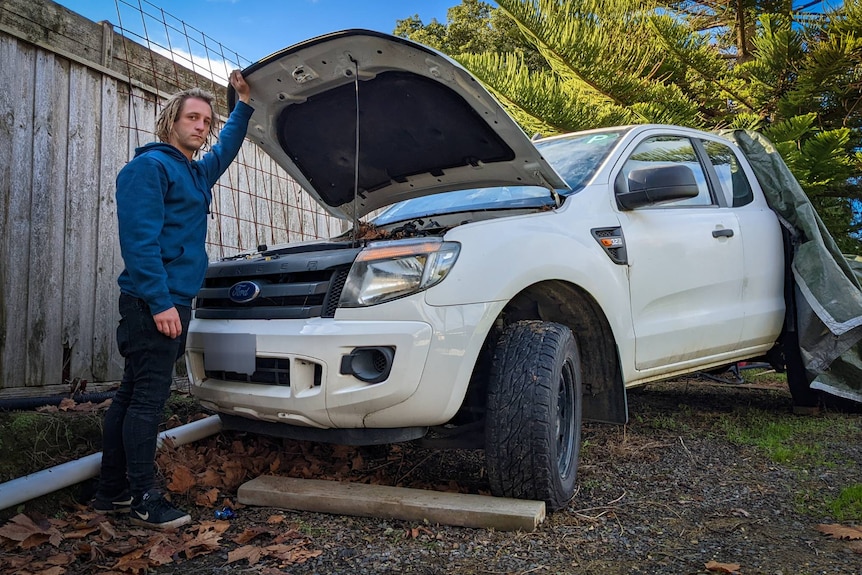A young man in his twenties looks at the camera, holding up the bonnet of the white ute, a tarp is over the back.