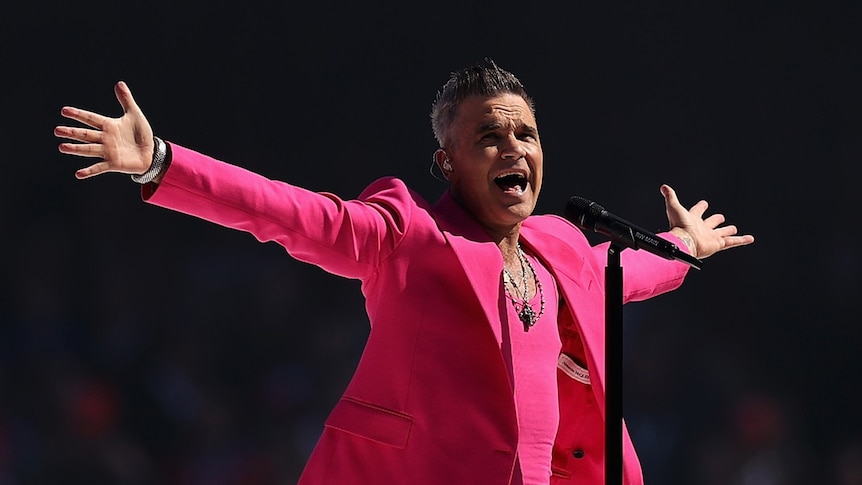 Robbie Williams spreads his arms as he stands on stage in a bright pink suit.