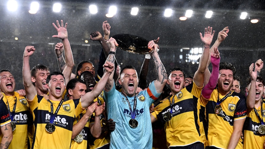 A professional soccer team lift a trophy, in front of a sign that reads premiers