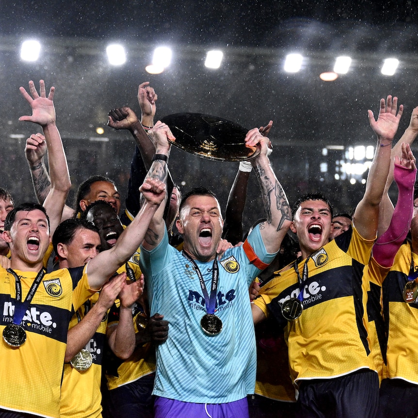 A professional soccer team lift a trophy, in front of a sign that reads premiers