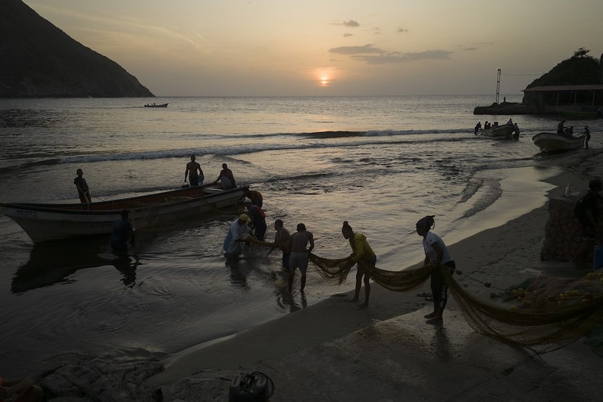A group of people pull a fishing net onto the beach from their boat at dusk