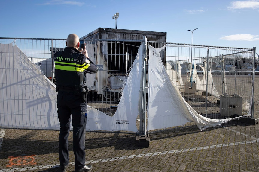 A police officer holds up a mobile phone to take a photo of a burned building, which is behind a fence.